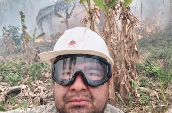 Seorang pria dengan menggunakan helm dan kaca mata pelindung memandang ke kamera, sementara di belakangnya semak-semak terbakar dan asap mengepul