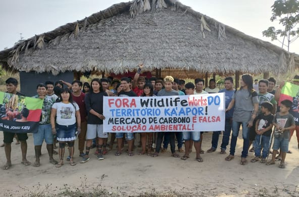 Sebuah kelompok terdiri dari 30 anggota masyarakat adat di depan pondok yang beratapkan pelepah kelapa melakukan protes dengan memegang spanduk: "Wildlife Works keluar dari wilayah Ka’apor. Perdagangan karbon adalah pemecahan krisis lingkungan"