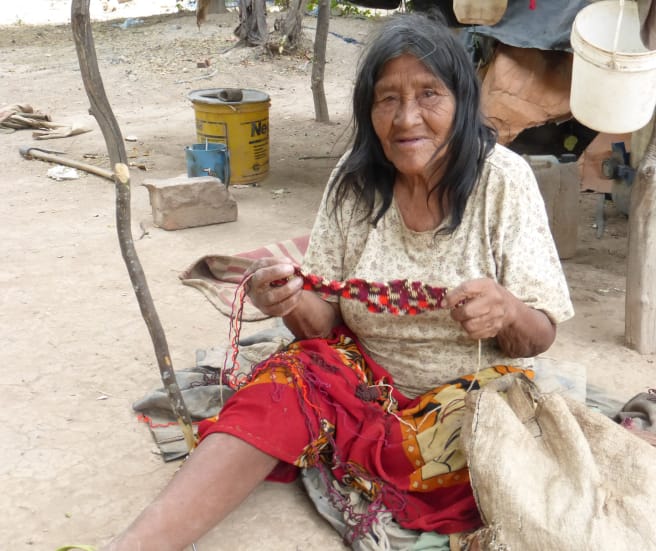 Seorang perempuan masyarakat adat yang duduk di atas tanah membuat rajutan dari serat tumbuhan.