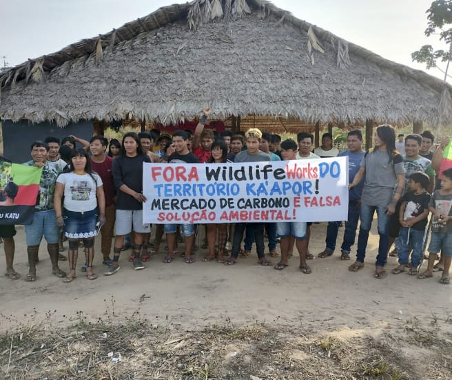 Sebuah kelompok terdiri dari 30 anggota masyarakat adat di depan pondok yang beratapkan pelepah kelapa melakukan protes dengan memegang spanduk: "Wildlife Works keluar dari wilayah Ka’apor. Perdagangan karbon adalah pemecahan krisis lingkungan"