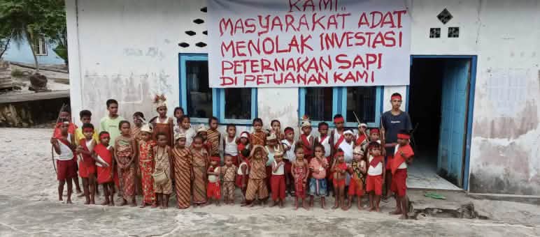 Anak-anak di depan sekolahnya dengan spanduk: „Kami masyarakat adat menolak investasi peternakan sapi di petuanan kami“