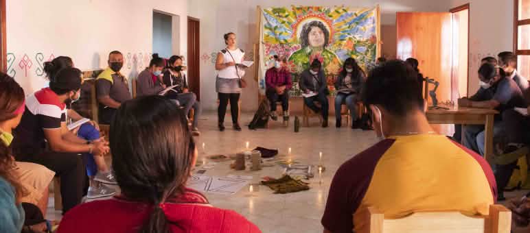 Para peserta duduk berlingkar. Mereka mengikuti sebuah workshop. Di tengah-tengah berdiri lilin yang menyala dan tergeletak kertas yang bertuliskan PAZ. Di dinding tergantung sebuah lukisan wajah Berta Cáceres.