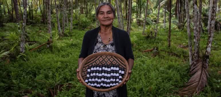 Seorang perempuan masyarakat adat yang berada di dalam hutan hujan sedang mempertunjukkan sebuah keranjang yang penuh dengan botol-botol kecil berisikan minyak eteris