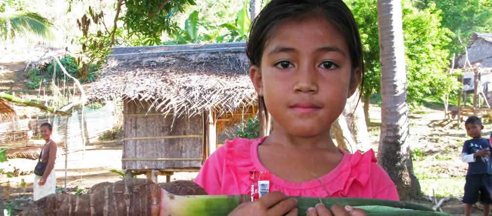 Seorang anak gadis sedang memangkul setumpukan umbi talas. Latar belakang nampak beberapa pondok