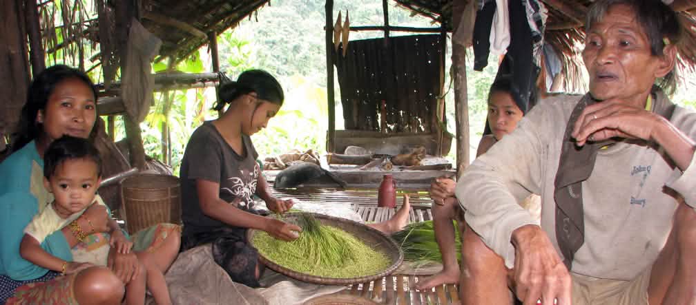 Seorang laki-laki, tiga perempuan dan satu anak kecil sedang duduk di pondok, sementara seorang perempuan sedang menampi beras