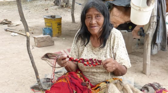 Seorang perempuan masyarakat adat yang duduk di atas tanah membuat rajutan dari serat tumbuhan.
