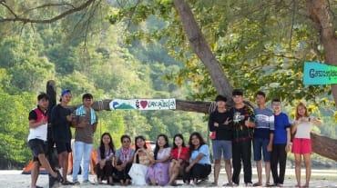 Sebuah kelompok anak muda sedang duduk dan berdiri di depan pohon di pantai