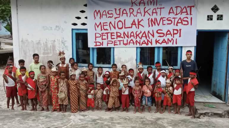 „Kami masyarakat adat menolak investasi industri ternak sapi di tanah kami!“ Anak-anak di depan sekolahnya dengan spanduk: „Kami masyarakat adat menolak investasi peternakan sapi di petuanan kami“