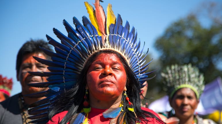 Seorang perempuan adat dihiasi dengan mahkota bulu burung di depan dua warga adat lainnya