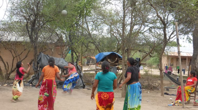 Lima perempuan masyarakat adat sedang bermain bola tangan disekitar pepohonan dan rumah kayu