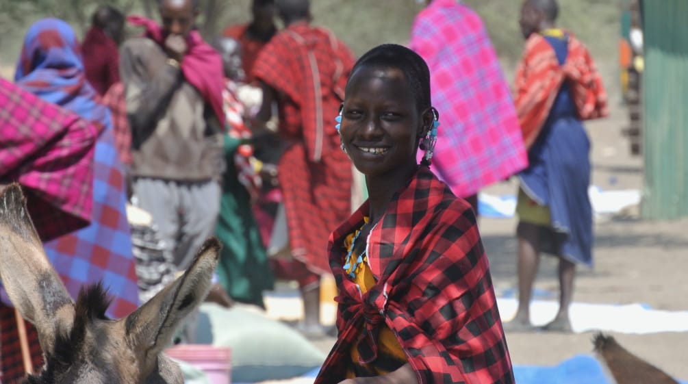 Perempuan dan laki-laki suku Maasai dengan sarung berwarna-warni di pasar, seorang perempuan menatap kamera
